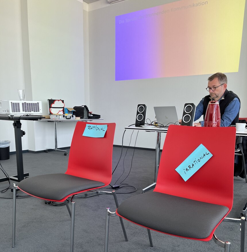 Zwei rote Stühle im Vordergrund und Martin Ziegenhagen ein Mann mit Brille sitzt im Hintergrund an einem Tisch