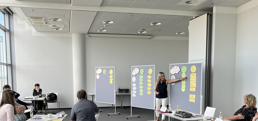 Mehrere Teilnehmende der Veranstaltung sitzen an Tischen und hören der Referentin zu. Diese hat blonde Haare und erklärt gerade etwas an einem Flipchart.