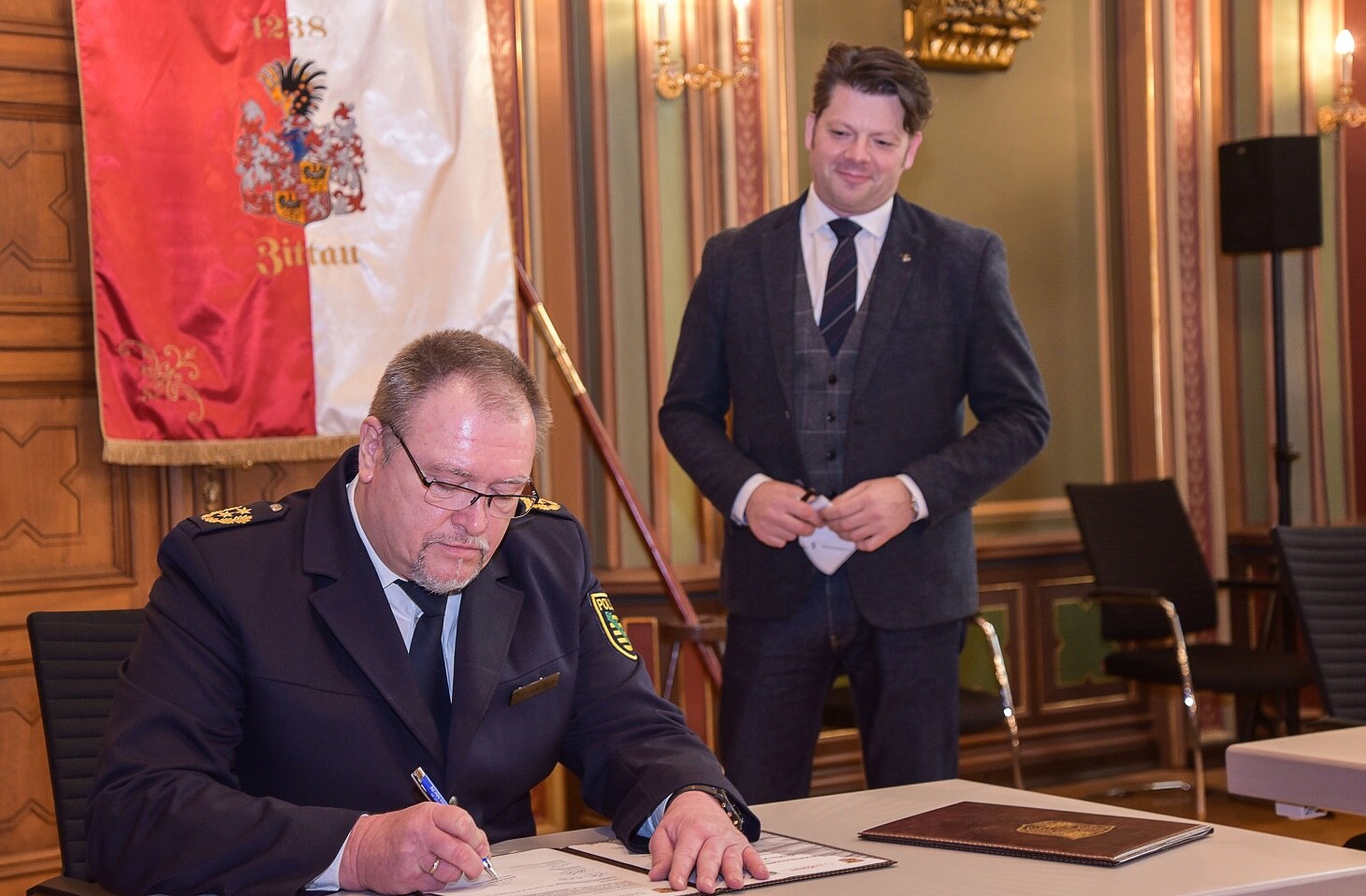 Herr Polizeipräsident Weißbach sitzt am Tisch und unterschreibt die Kooperationsvereinbarung. Herr Oberbürgermeister Zenker steht daneben und beobachtet ihn dabei.