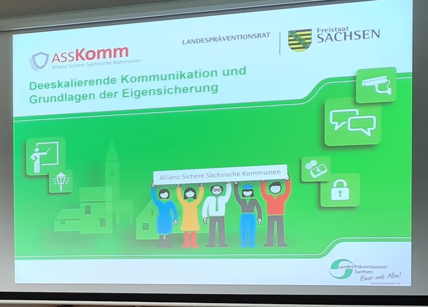 Präsentation des Vortragen von Torsten Kosuch zum Thema Gewalt und dessen Umgang beim Seminar Deeskalationstraining. Man sieht eine digitale Leinwand mit einer grünen Folie auf der das typische ASSKomm Logo zu sehen ist und dann das Thema des Vortrages