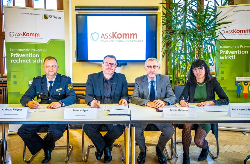 Die drei Unterzeichnenden sitzen mit dem Innenminister an einem Tisch. Im Hintergrund sieht man zwei Roll up's mit dem ASSKomm-Logo.