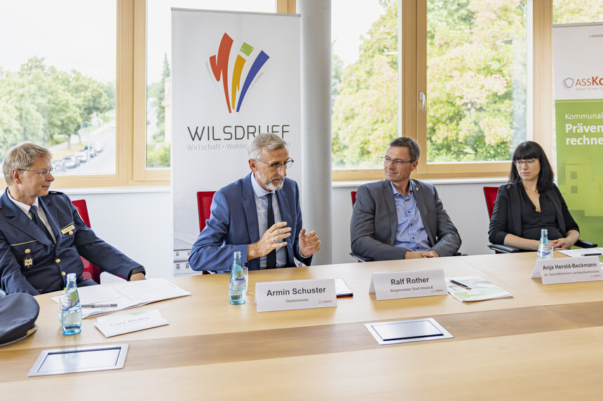 Vier Personen sitzen an einem Tisch auf dem Unterlagen liegen. Hinter ihnen steht ein Roll up mit dem Logo von Wilsdruff und ein Roll up mit ASSKomm-Logo.