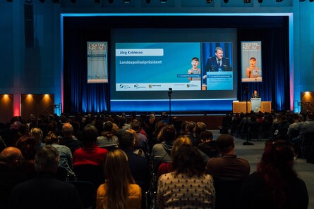 Der Landespolizeipräsident steht auf der Bühne zur Eröffnungsrede und wird auf die große Leinwand übertragen.