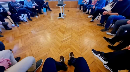 Die Schuhe der Teilnehmenden, aus der Vogelperspektive fotografiert, darunter warmer Parkettboden. Sie sitzen auf Stühlen im Kreis zusammen.