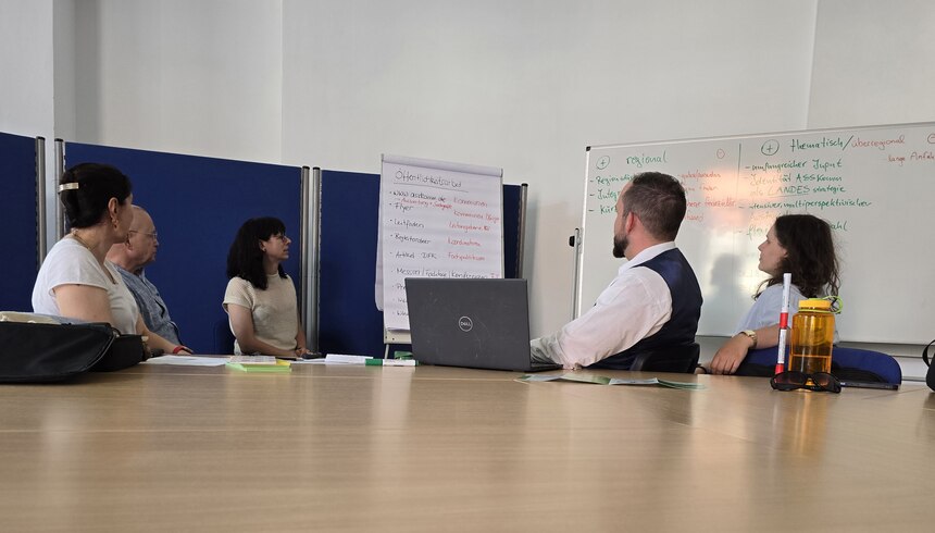 Fünf Personen sitzen mit dem Rücken zum Fotografen und schauen auf ein Whiteboard.