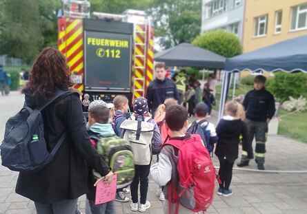 Das Bild zeigt eine kleine Gruppe von Kindern vor einem Feuerwehrwagen. Ihnen stehen zwei Feuerwehrmänner gegenüber.