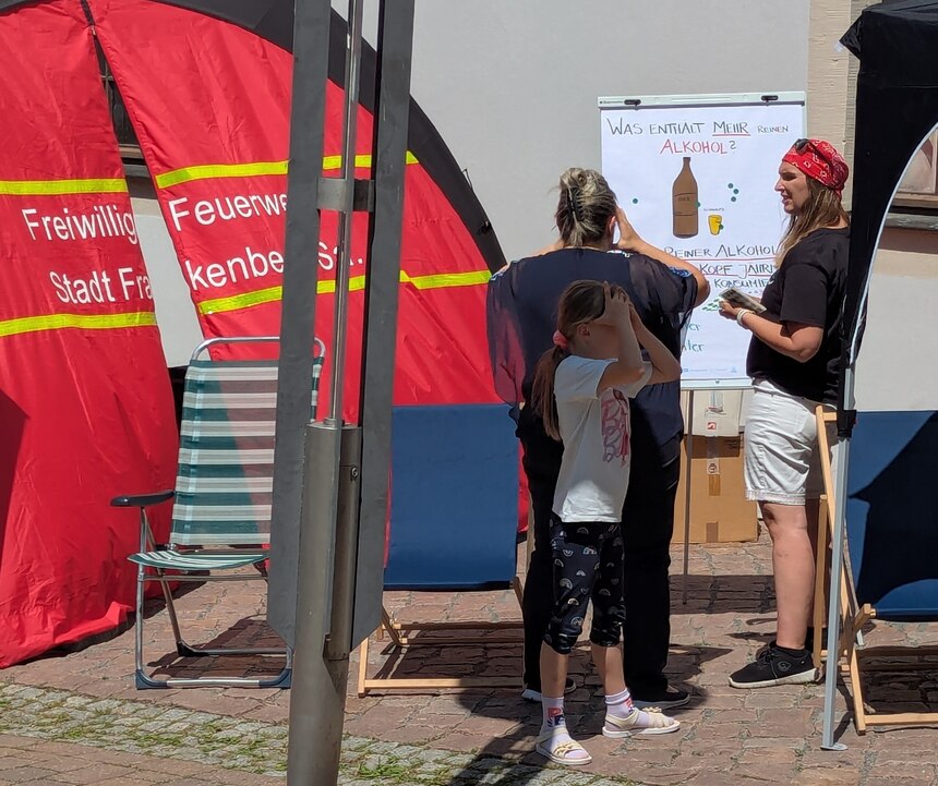 Zu sehen ist ein rotes Feuerwehrzelt mit einer kleinen Gruppe Menschen davor. Daneben steht ein Flipchart mit der Aufschrift »Was enthält mehr reinen Alkohol?« .