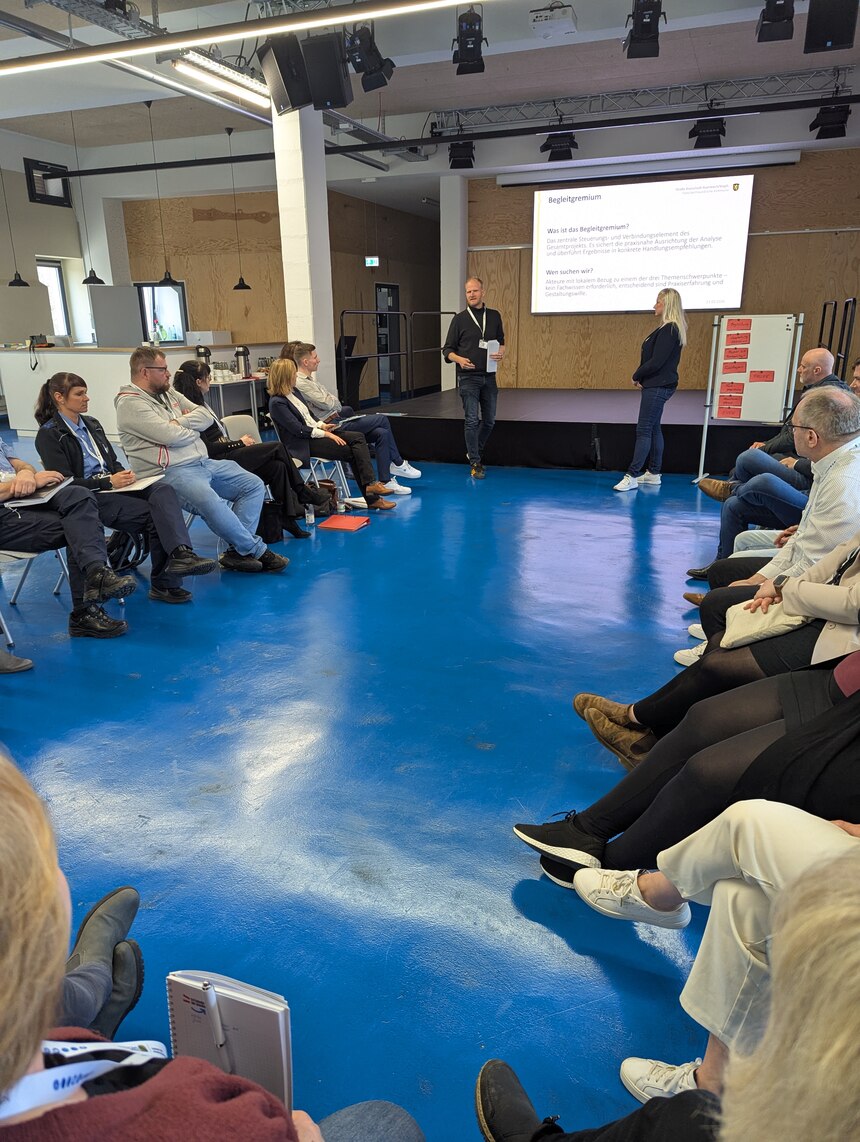 Die Teilnehmenden sitzen in einem Kreis. Vorn sieht man eine Präsentation und ein Mann steht links davon und eine Frau rechts.
