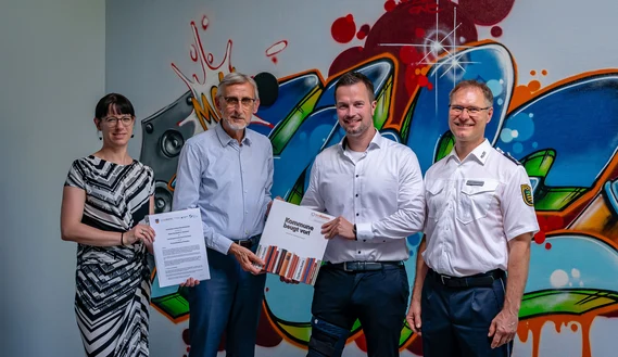 Vier Personen stehen vor einer Graffiti-Wand und halten die Kooperationsvereinbarung hoch.