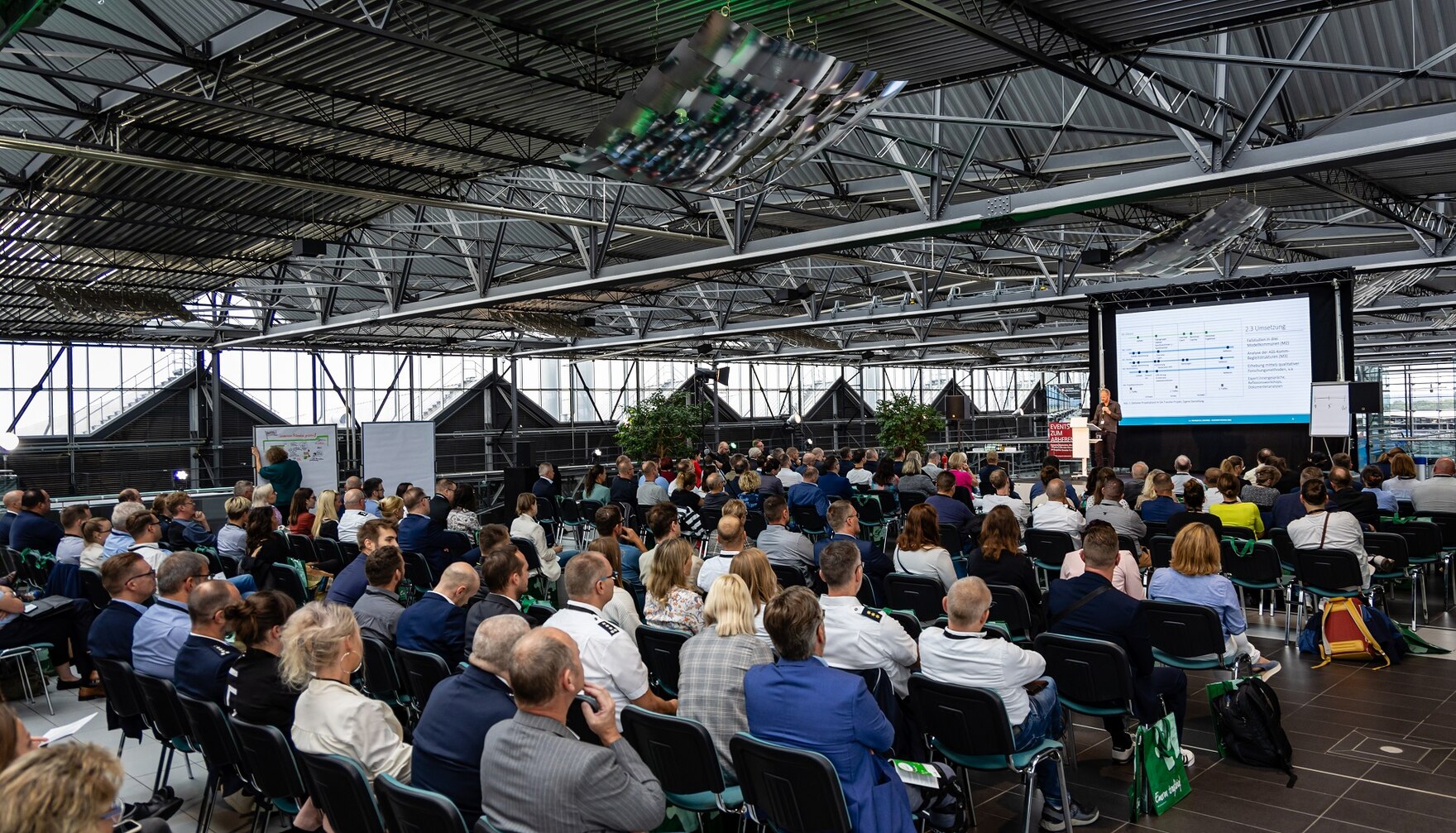 Plenum des Fachtages von hinten mit Teilnehmern fotografiert. Auf der Bühne steht als Referent Dr. Jan Lorenz Wilhelm. Im Hintergrund sieht man seine Präsentation.