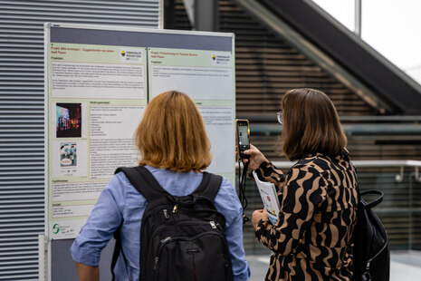 Zwei Personen stehen vor zwei Plakaten. Eine Person fotografiert ein Plakat.