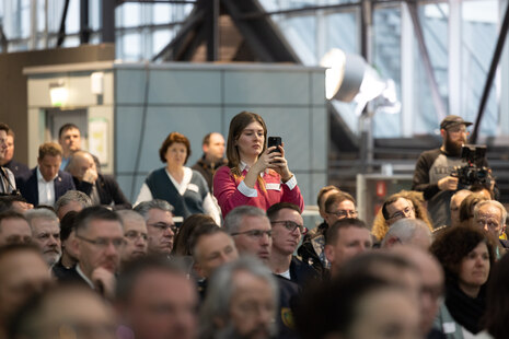 Blick ins sitzende Publikum. Eine Frau steht und hält ihr Handy hoch.