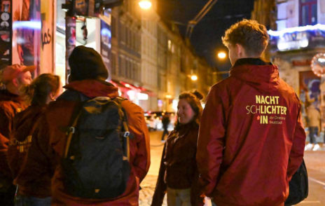 Nachtschlichter*innen steht in den mit dem Logo bedruckten Jacken am Abend auf einer Straße in der Dresdner Neustadt