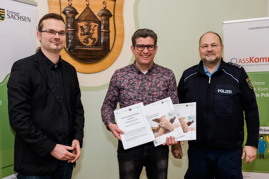 der Geschäftsführer Landespräventionsrat Sven Forkert, Bürgermeister Lars Naumann sowie der Leiter Polizeirevier Rochlitz Jens Rödel stehen nebeneinander. Bürgermeister Lars Naumann hält die unterzeichnete Kooperationsvereinbarung in den Händen. 