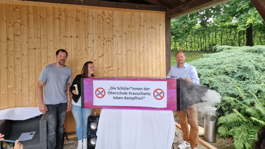 drei Personen bei der Präsentation ihres Projektes, sie stehen neben einer Nachempfunden E-Zigaretten mit der Aufschrift: Die Schülerinnen der Oberschule Krauschwitz leben dampffrei!