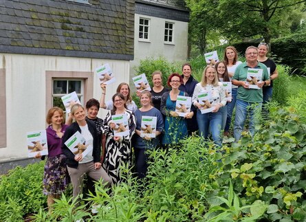 Im Hintergrund sieht man eine helle Hausfassade und im Vordergrund grüne Pflanzen. In der Mitte befinden sich in einer Diagonalen 14 Personen. Dabei halten die 13 Frauen und ein Mann eine helle Mappe in der Hand und lächeln.