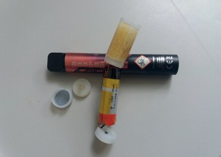 Die einzelnen Bestandteile einer E-Zigarette bzw. Vape liegen in Vogelperspektive auseinandergebaut auf einem Tisch. Ein Totenkopfsymbol ist auf der Plastikhülle. Außerdem liegt eine zylinderförmige Batterie auf dem Tisch.