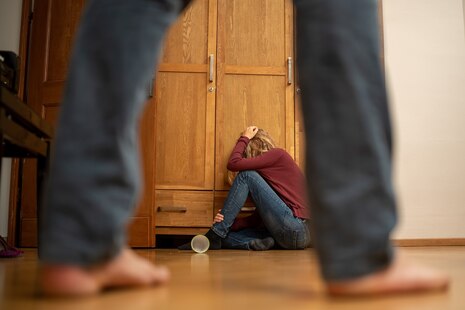 Ein Mädchen kauert mit den Händen schützend über dem Kopf vor einem Schrank und man sieht noch zwei Männerbeine.