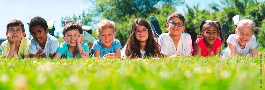 Mehrere Kinder liegen bäuclinks auf einer Wiese und lachen in die Kamera. Es sind 6 Kinder, 5 Jungs und ein Mädchen