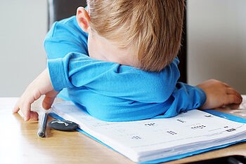 Ein kleiner Junge ist über seinen Hausaufgaben gebeugt, mit dem Kopf in seiner Armbeuge.