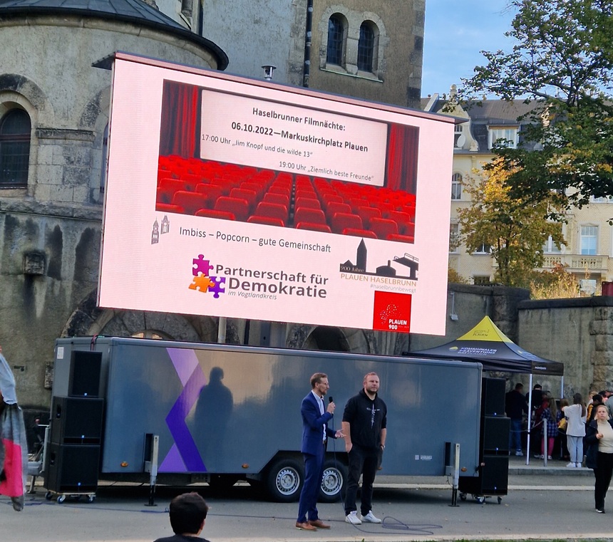 Bild zeigt Kulturbürgermeister der Stadt Plauen Tobias Kämpf sowie eine weiter Person bei der Eröffnung der Veranstaltung vor der Leinwand