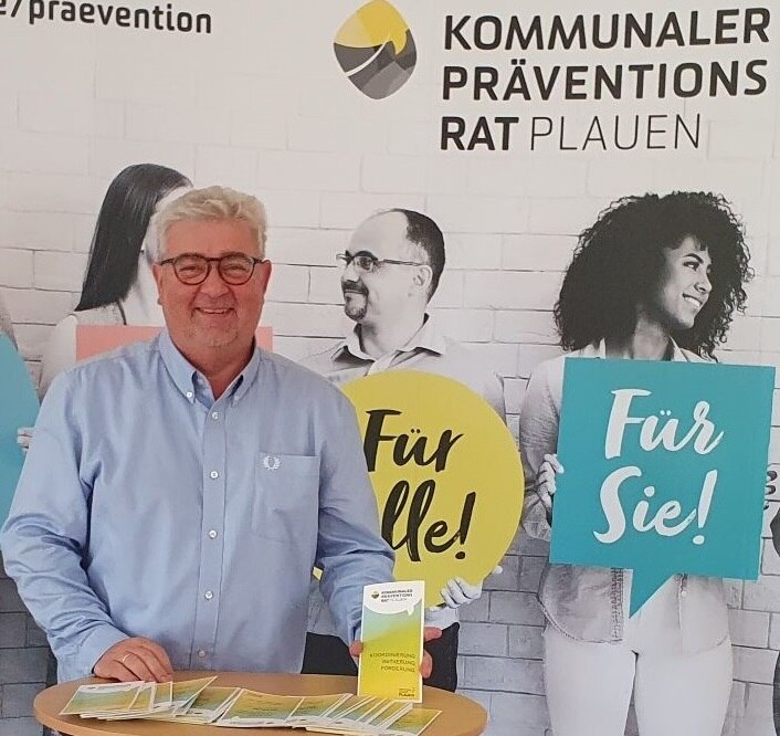 Mann mit weißen Haaren, einem hellblauem Hemd steht an einem Infostand