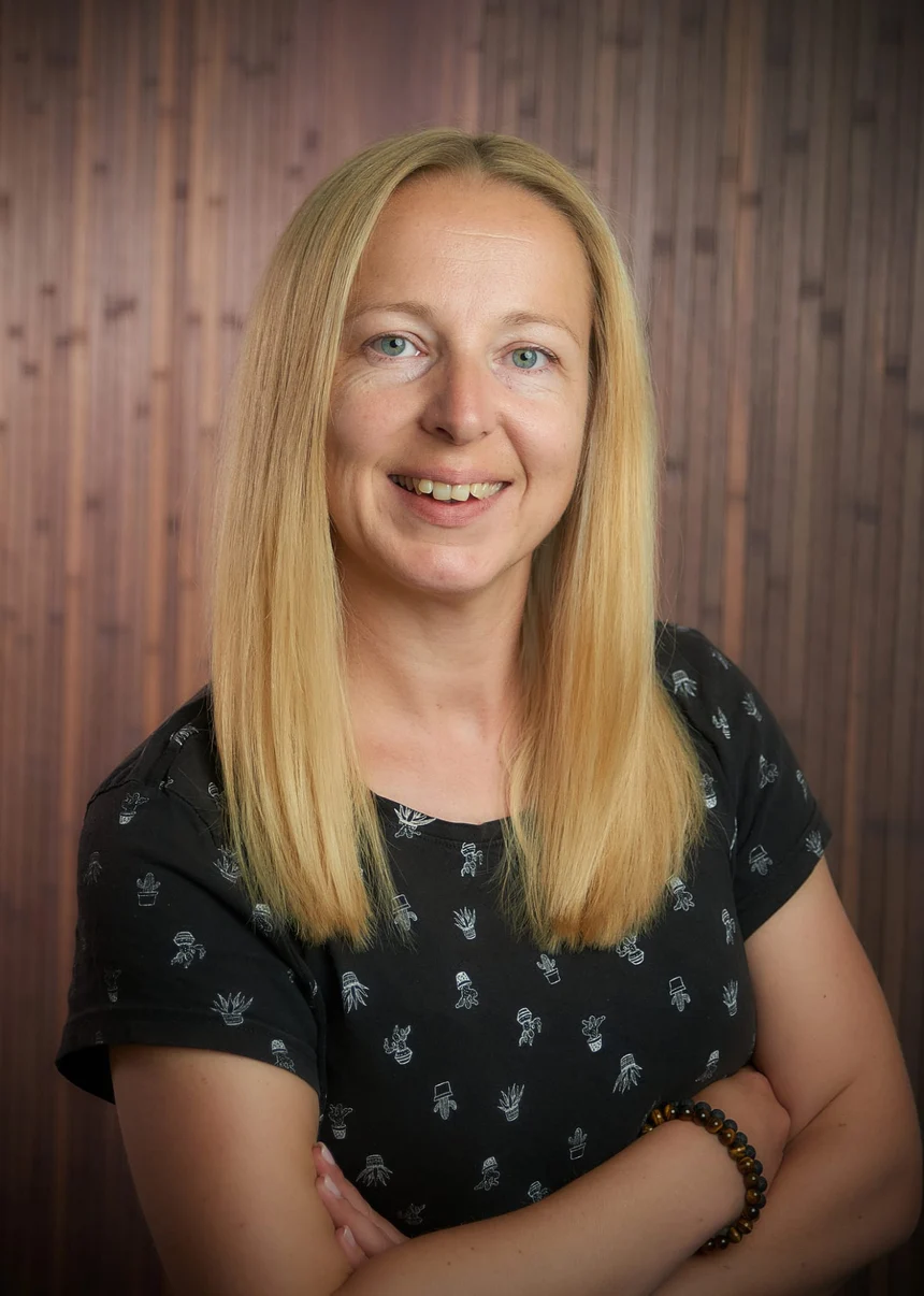 Portrait von Monique Weixler. Sie trägt die langen blonden Haare offen, hinter ihr eine dunkle Holzwand.