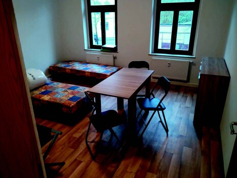 Auf dem Bild sieht man ein Zimmer mit zwei Betten, einem Tisch, drei Stühlen und einer Kommode.