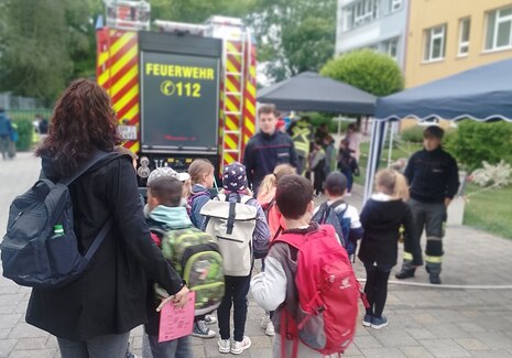 Kinder stehen vor einer Schule und betrachten ein Feuerwehrauto und zwei Feuerwehrmänner.