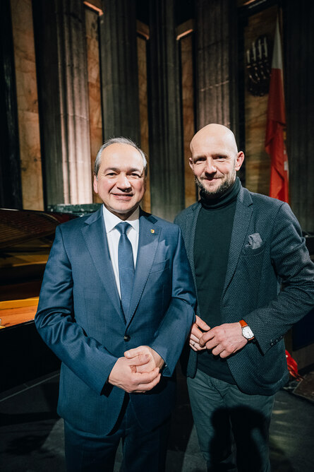Zwei Männer stehen vor einem dunklen Hintergrund und lächeln ins Bild.