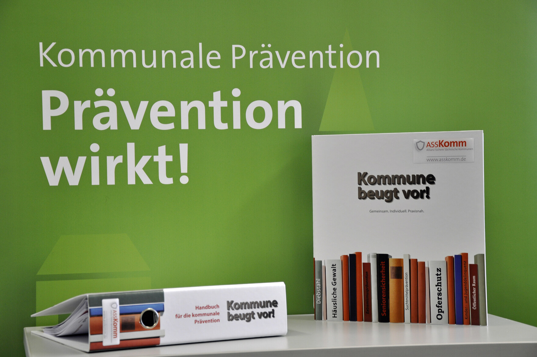 Vor dem grünen ASSKomm Hintergrund mit dem Schriftzug "Prävention wirkt" stehen die AsskommOrdner nebeneinander. Der eine mit dem Ordnerrücken zur Kamera, der andere mit der Vorderseite zur Kamera.