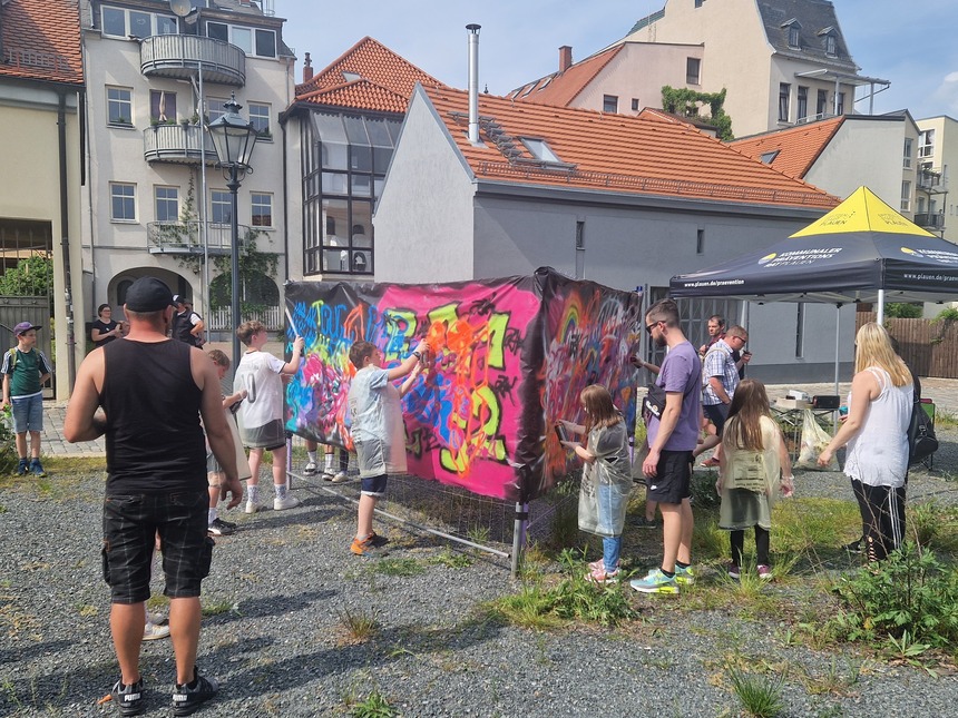 zeigt Kinder und Eltern welche an eine Plane Graffiti sprühen, im Hintergrund ein Schwarzes Festzelt des KPR Plauen