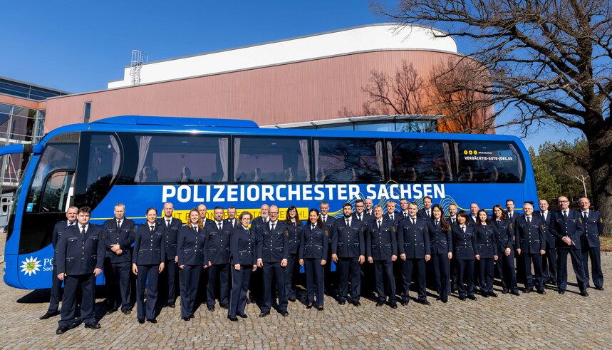 zeigt die Mitglieder des Polizeiorchesters Sachsens in Uniform vor einem Reisebus mit der Aufschrift Polizeiorchester Sachsen