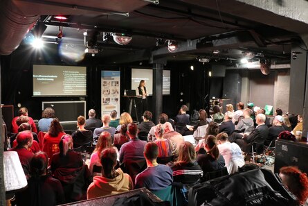 Blick zur Bühne mit einer Referentin und das Publikum.