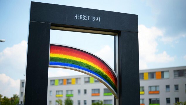 Zu erkennen ist ein Denkmal mit einem bunten Regenborgen und der Überschrift »Herbst 1991« in schwarz grau. 