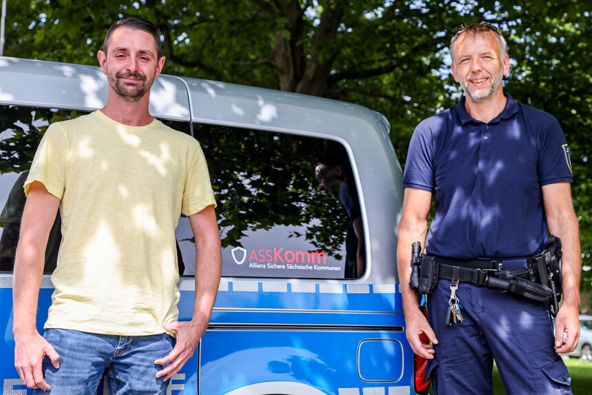 Zwei Männer lehnen an einem Polizeiwagen, der eine der rechts steht ist in Polizeiuniform gekleidet, der andere hat ein gelbes Tshirt und eine Jeans an