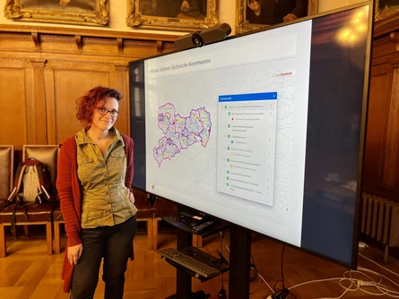 Maria J. Schubert steht vor einer Präsentationstafel mit einer Karte von Sachsen im Ratssaal des Rathauses Görlitz..