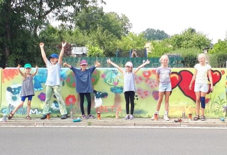 Kinder recken die Arme in die Höhe und stehen vor ihrem selbst gestalteten Graffiti.