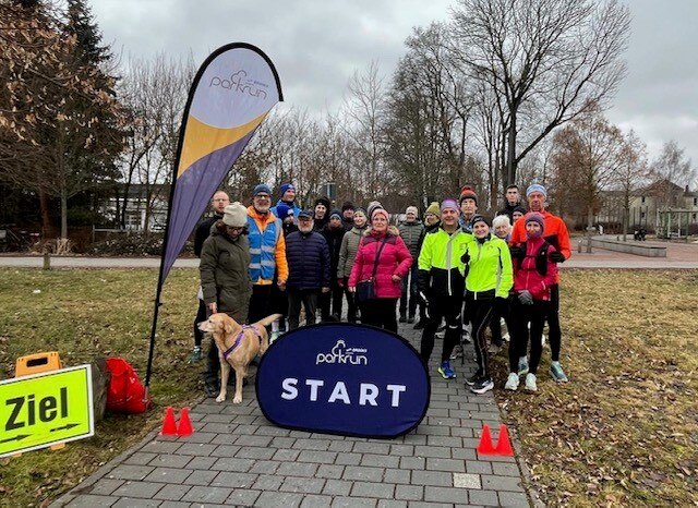 21 Personen und ein Hund stehen auf dem Rasen. Daneben steht eine Fahne mit dem Aufdruck "parkrun".