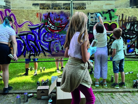 Im Hintergrund sieht man mehrere Personen an einer Wand mit Graffiti. Im Vordergrund ist ein Kind mit einer Sprühdose.